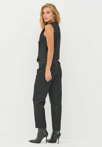 Zwarte vest met textuurpatroon, gecombineerd met hoge zwarte broeken. Model draagt gestudde zwarte hoge hakken.