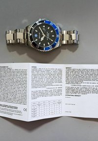 Orologio da polso con bracciale in acciaio inossidabile, quadrante nero e lunetta blu. Include garanzia e manuale di istruzioni, che mostra vari dati tabellari.