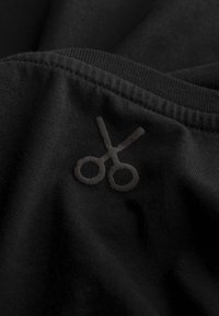 Zwarte stof met een getextureerde, geborduurde schaar in een iets donkerdere tint zwart, gepositioneerd aan de rand van het materiaal.