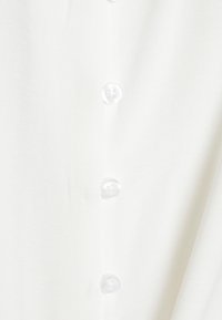 Camicia bianca con bottoni sul davanti, realizzata in un tessuto leggero e liscio con una superficie leggermente strutturata.