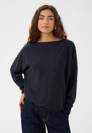 Une femme porte un sweat-shirt noir ample à encolure bateau et un jean foncé sur un fond clair uni.