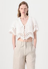 Hvit kort bluse med bølget kant og øyelet detaljer, med en knyteåpning foran og vide ermer. Sammen med beige bukser.