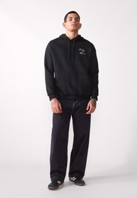 Sudadera negra con bolsillo frontal y cordones, con texto bordado. Combinada con jeans de mezclilla oscuros y zapatillas negras. Textura suave.