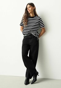 Črno-bela črtasta majica s sproščenim krojem, kombinirana z visokim pasom črnih kavbojk, dopolnjena s črnimi čevlji z zašiljenimi konicami.