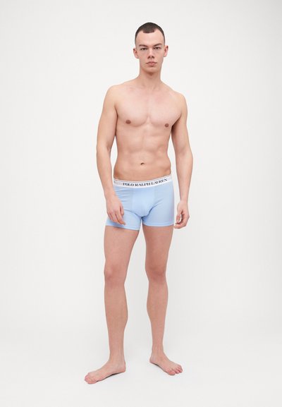 Boxeri scurți de culoare albastră deschisă cu un brau alb pe care se află logo-ul "POLO RALPH LAUREN". Fabricată dintr-un material neted și elastic. Modelul stă desculț.