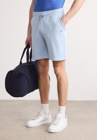 Homme portant un short bleu clair à cordon, des baskets blanches et des chaussettes blanches, tenant un sac de sport bleu marine à ses côtés.