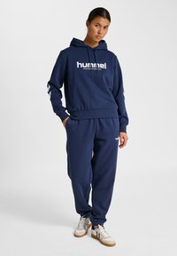 Junge Frau, die in einem navyblauen Hummel-Hoodie und passender Jogginghose mit weißen Turnschuhen vor einem weißen Hintergrund steht.