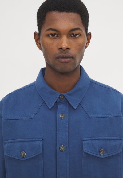 Camicia di cotone blu con chiusura a bottoni, colletto strutturato, due tasche sul petto e bottoni a contrasto. Tessuto liscio con un colore intenso.