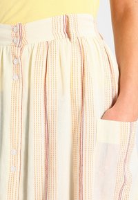 Gonna a righe gialla realizzata in un tessuto leggero con linee verticali, bottoni e una tasca laterale. I colori sono tenui con sottili accenti rossi.