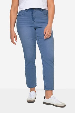 Person iført lyseblå slim-fit jeans, hvide sneakers med beige og sorte detaljer og en hvid top, står med armene afslappede.