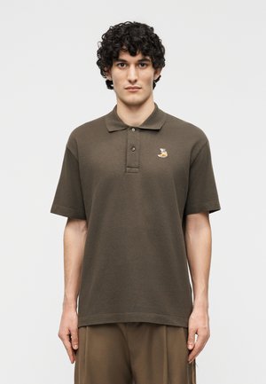 Jeune homme aux cheveux bouclés portant un polo à manches courtes marron avec un petit logo de renard sur la poitrine gauche.