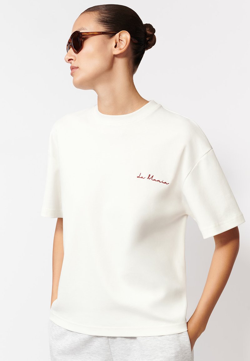 Biała bawełniana koszulka z krótkim rękawem i okrągłym dekoltem; zdobiona małym, czerwonym haftowanym napisem "la Marais". Gładka tekstura, luźny krój.