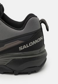 Czarno-szare buty trailowe z siateczkową cholewką, teksturowanymi akcentami, wzmocnionym obcasem i wyraźnym logo "SALOMON" po bokach.