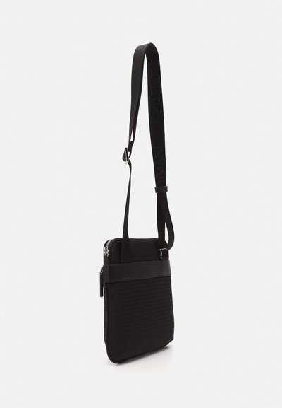 Black crossbody bag made of textured fabric with a smooth leather accent, featuring a zip closure and adjustable strap with branding.