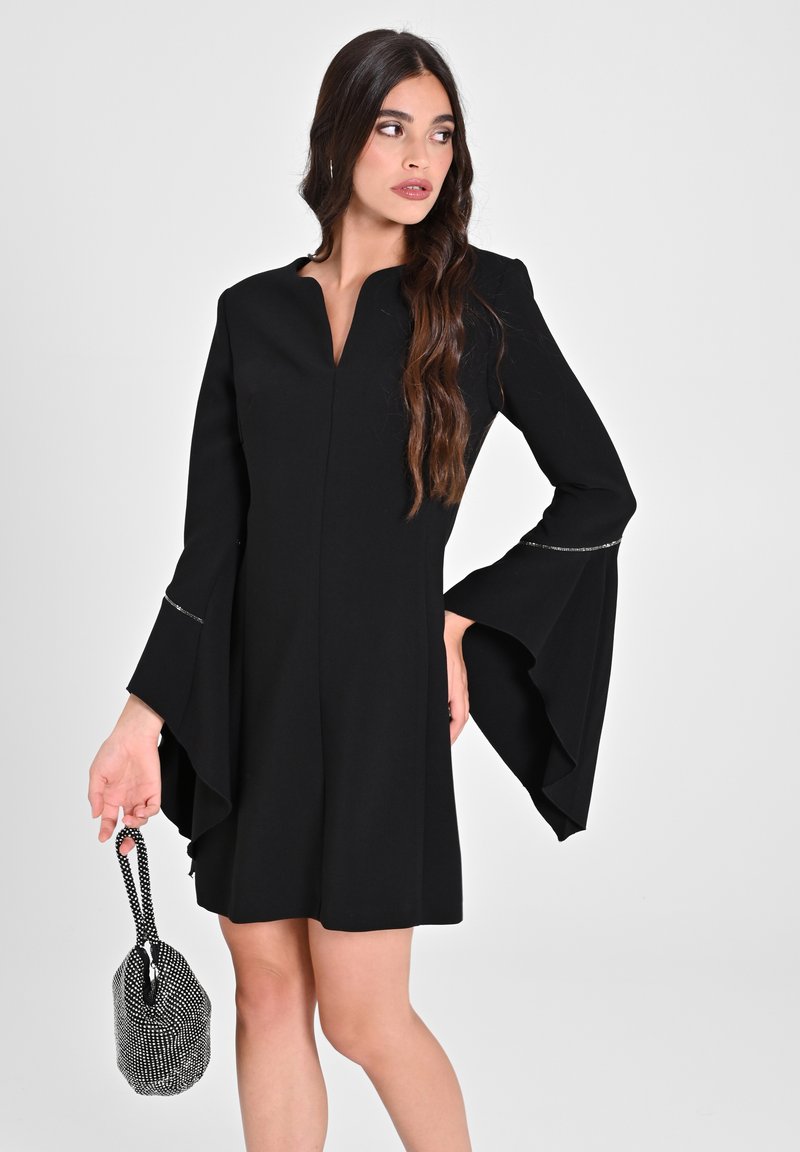 Schwarzes Kleid mit langen Glockenärmeln, V-Ausschnitt und silbernen Akzenten an den Ärmeln. Das Modell hält eine kleine schwarz-weiße Handtasche.