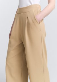Pantaloni beige su misura con vita alta, dotati di tasche laterali e una morbida texture del tessuto. Pieghe che scendono davanti per una forma strutturata.