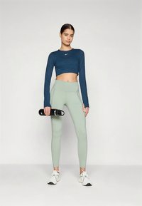 Blaues, kurz geschnittenes Langarmoberteil und hellgrüne, hoch taillierte Leggings. Model hält eine schwarze Wasserflasche. Weiße Sportschuhe vervollständigen das Outfit.