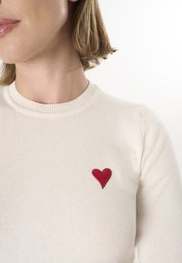 Crèmekleurige trui met een ronde hals, met een rode geborduurde hart op de linkerkant. De stof lijkt zacht en gestructureerd.