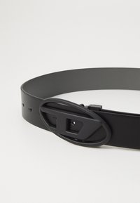 Cinturón de cuero negro con un acabado suave, que presenta una gran hebilla ovalada en un tono negro mate y un cierre de una sola pua. Longitud ajustable.