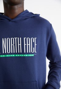 Mörkblå hupptröja med stor vit logotyp "THE NORTH FACE" och en grön detalj under med texten "NEVER STOP EXPLORING."