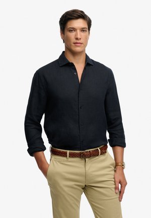 Ung mand iført en sort skjorte med knapper og op rullede ærmer, beige bukser, brunt flettet bælte og et armbåndsur, hænder delvist i lommerne.