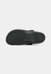 Chaussure Crocs noire vue par-dessous, affichant une semelle texturée avec des motifs d’adhérence rectangulaires et en forme de chevrons.