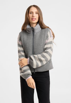 Woman with long hair wearing a gray zip-up vest over a gray and white striped sweater with pink cuffs, standing with arms crossed.