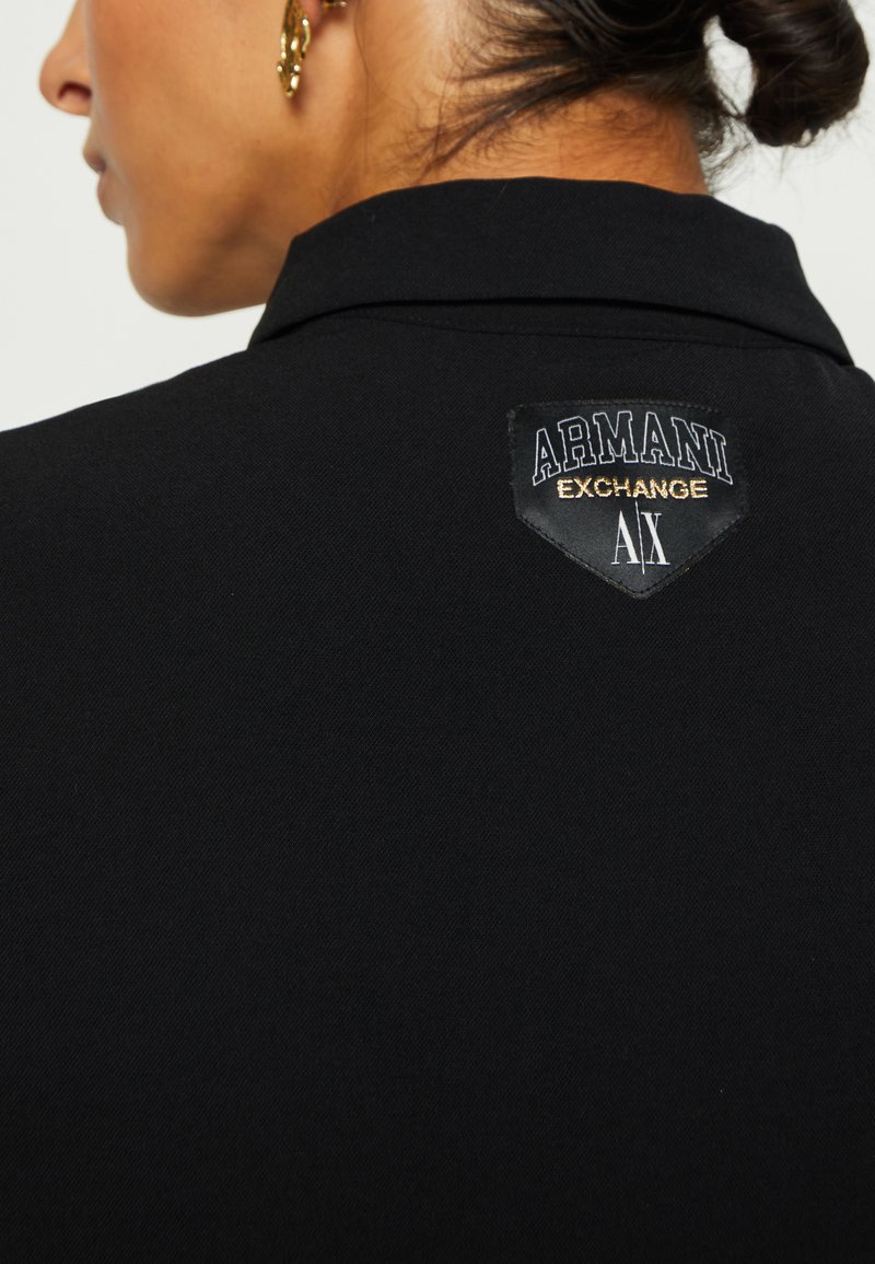 Schwarzes Poloshirt mit strukturiertem Finish, versehen mit einem Aufnäher, der "Armani Exchange" in Weiß und Gold zeigt. Der Kragen hat ein traditionelles Design.