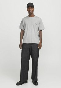 Camiseta de algodón gris con manga corta, que presenta un logo impreso. Combinada con pantalones vaqueros negros de corte holgado y botas negras.