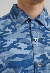 Denim ing kék és világoskék terepmintával, hegyes gallérral, gombos elülső résszel és fémgombokkal.