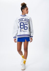 Szary, oversize'owy sweatshirt z niebieskimi paskami i graficznym napisem; zestawiony z niebieską mini spódnicą, żółtymi skarpetkami w paski oraz białymi sneakersami.