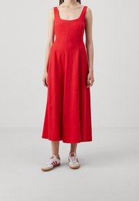 Robe en lin rouge avec un décolleté carré et de larges bretelles, évasée à partir de la taille jusqu'à mi-mollet, associée à des baskets blanches.