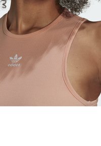 Męska bezrękawnikowa, żebrowana koszulka sportowa w jasnym brzoskwiniowym kolorze, z okrągłym dekoltem i małym, metaliczno-srebrnym logo Adidas na piersi.