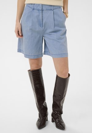 Kvinde iført lyseblå cowboyshorts med forstykker foran og høje mørkebrune knæhøje støvler med krokodillemønster, hænder delvist i lommerne.
