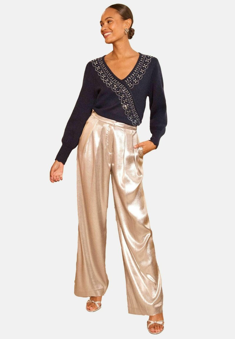 Jūras spēku svīteri ar rotātu apkakli, apvienojot ar platu kāju, spīdīgām bežām biksēm. Modelis stāv ar vienu roku kabatā, demonstrējot elegantu siluetu.