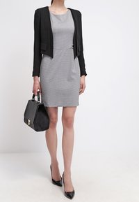 Robe à carreaux gris au silhouette cintrée, portée avec une veste noire courte et des escarpins noirs en vernis, tenant un sac à main structuré noir.