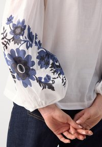 Manche de blouse blanche avec broderie florale bleue et noire, froncée au poignet, montrée avec les mains jointes derrière le dos et un jean en denim noir.