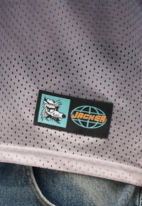 Δίχτυ από μαλακό λεβάντα, με μαύρη ετικέτα μάρκας που διαθέτει μπλε και λευκό γραφικό και πορτοκαλί κείμενο "JACKER." Φόντο από τζιν.