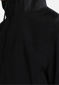 Sort vandtæt jakke med høj krave, lynlås foran og glat overflade. Har forstærkede syninger og minimalistisk design.