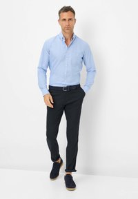 Lichtblauw overhemd met knoopsluiting en een borstzak, gecombineerd met donkere, slim-fit broek en marineblauwe sneakers met een gumrubberen zool, tegen een witte achtergrond.