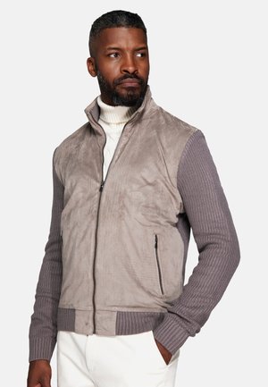 Man with beard wearing gray zip-up jacket with knitted sleeves and white pants, looking to the side with hand in pocket.