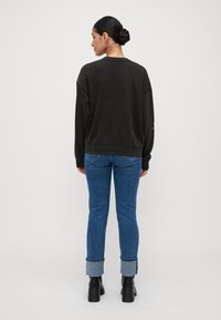 Haját bunban összefogó nő fekete pulóvert, kék, felhajtott szárú farmert és fekete, magas sarkú bokacsizmust visel, háttal állva egy egyszínű háttér előtt.