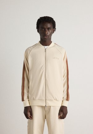 Krämfärgad zip-up jacka med ribbade muddar och nederkant, med röda och bruna ränder längs ärmarna. Slät textur, minimal branding ovanför dragkedjan.