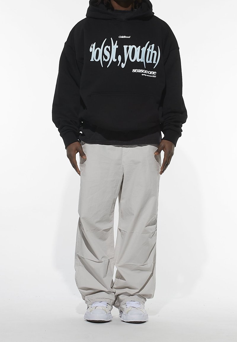 Sweat à capuche noir avec texte graphique en bleu ; porté avec un pantalon ample beige clair. Fond blanc uni. Les baskets sont blanches avec des accents.