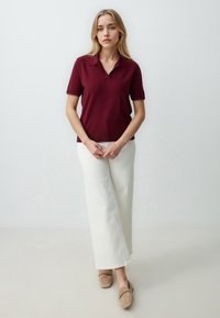 Burgundowa koszulka polo z krótkim rękawem, zestawiona z białymi spodniami o szerokich nogawkach i beżowymi butami wsuwanymi, na jasnym tle.