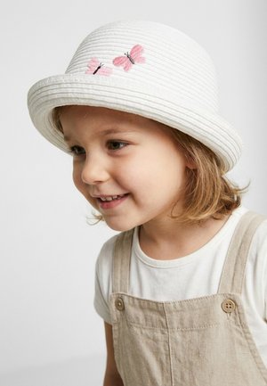 Junges Kind lächelt, trägt einen weißen Hut mit rosa Schmetterlingsstickerei und beige Latzhose über einem weißen Hemd, leicht nach links gewandt.