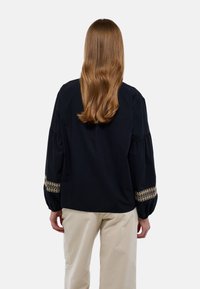 Blouse noire avec des manches bouffantes et des accents dorés brodés aux poignets. Portée avec un pantalon couleur crème, présentant une coupe et un design décontractés.