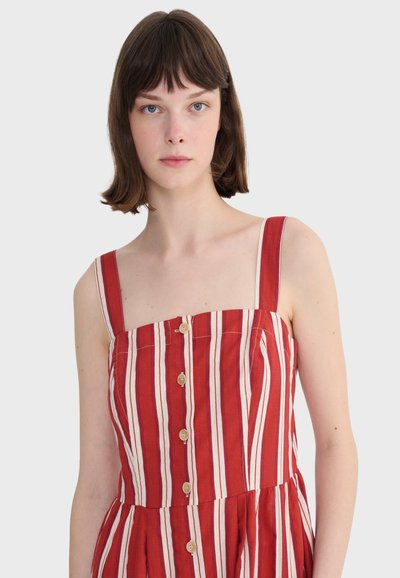 Jeune femme aux cheveux bruns courts portant une robe sans manches à rayures verticales rouges et blanches avec des boutons sur le devant.