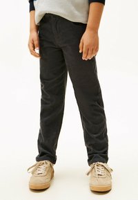 Kind in zwarte corduroy broek en beige sneakers, staand tegen een witte achtergrond, met handen ontspannen langs de zij.