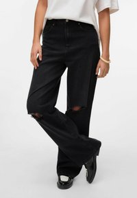 Zwarte spijkerbroek van denim met een relaxte pasvorm, voorzien van gescheurde knieën en subtiele stikseldetails, gecombineerd met glanzende zwarte schoenen.